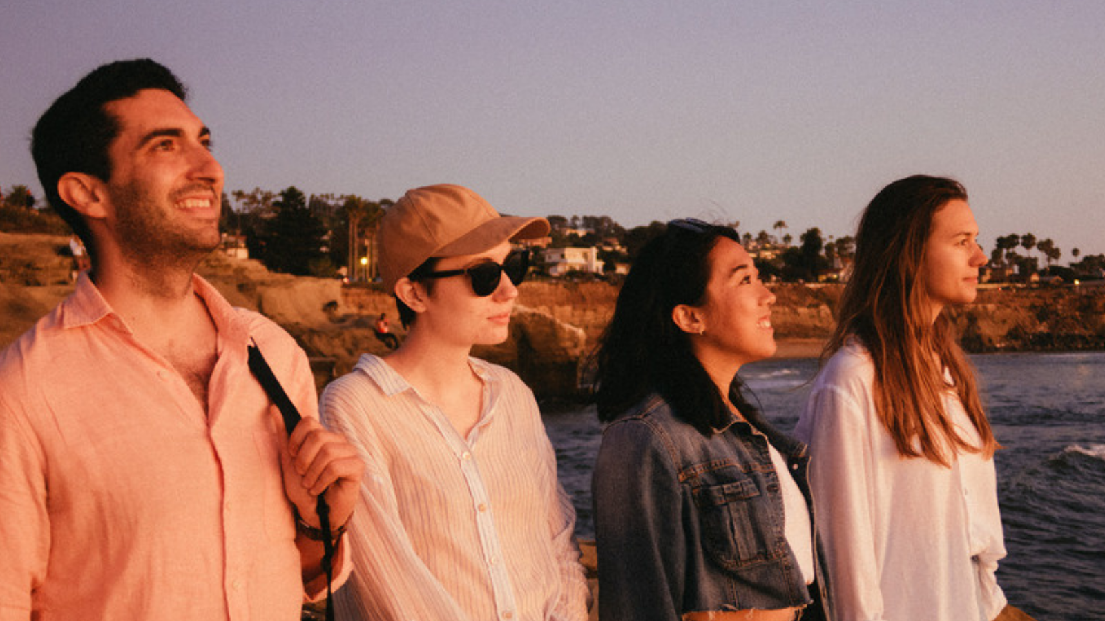 Four young people stand by the coast in evening light, looking in different directions