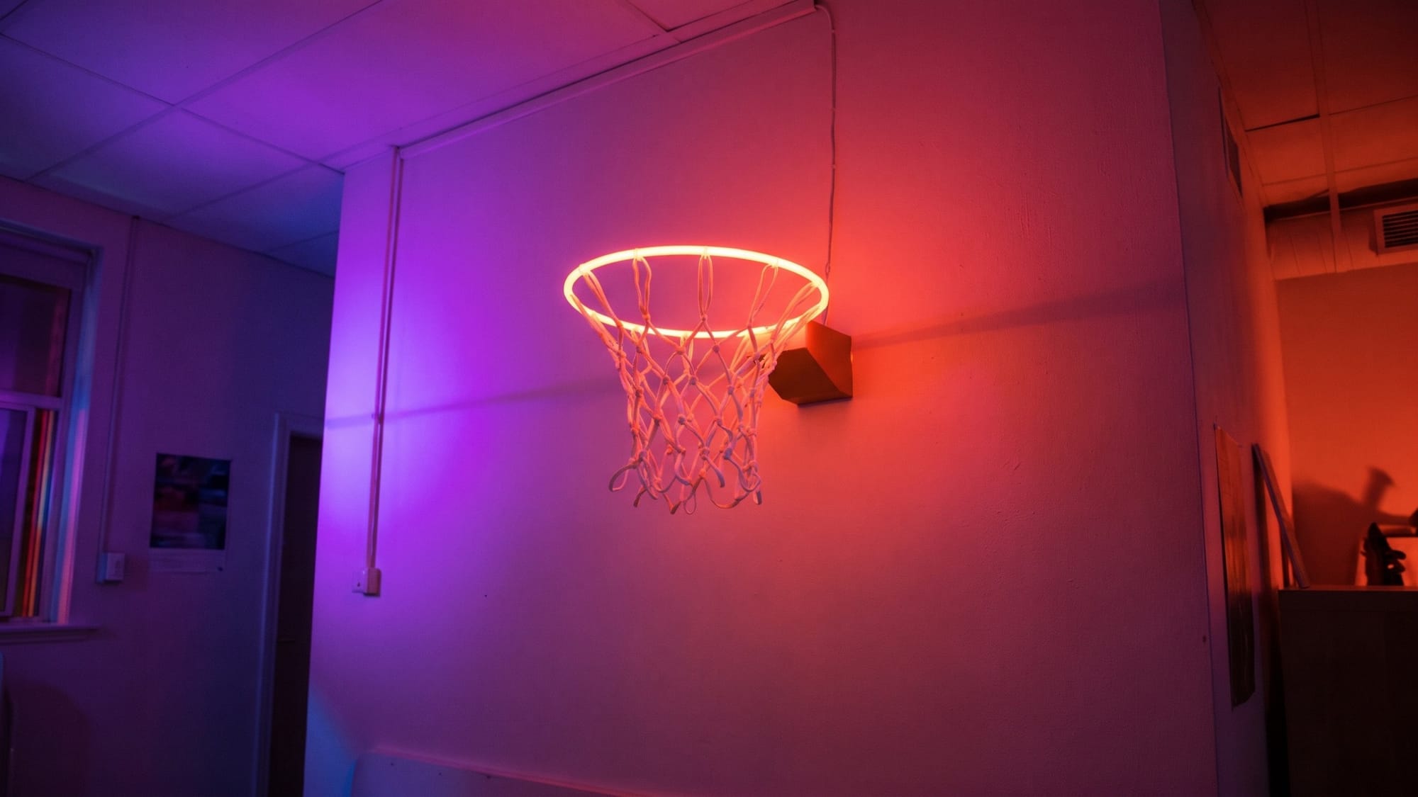 Svítící neonová basketbalová obroučka se síťkou na zdi prázdné místnosti v růžovém a fialovém světle.