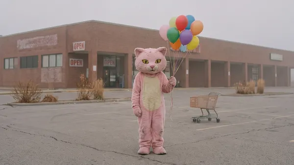 Maskot růžové kočky s barevnými balónky stojí sám na prázdném parkovišti u obchodu.