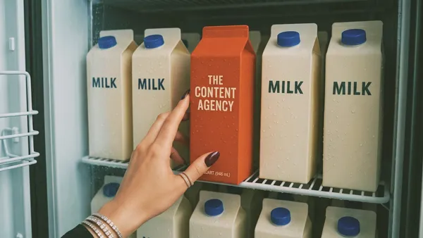 Ruka s prsteny bere z lednice oranžový karton s nápisem The Content Agency mezi bílými kartony mléka