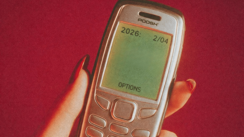 Ruka drží starý tlačítkový mobil s nápisem 2026 na displeji na červeném pozadí