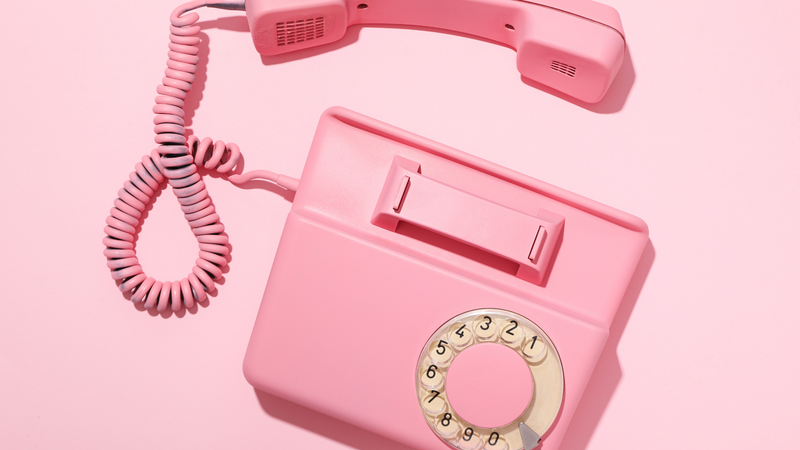 Růžový retro telefon s otočným číselníkem a vyvěšeným sluchátkem na růžovém pozadí