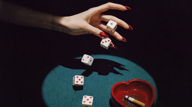 Ruka s rudými nehty hází bílé kostky se srdíčky na zelené sukno v kuželu světla, vedle stojí červený popelník ve tvaru srdce s cigaretou.