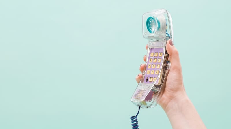 Ruka drží průhledné telefonní sluchátko se spirálovou šňůrou na mentolovém pozadí.